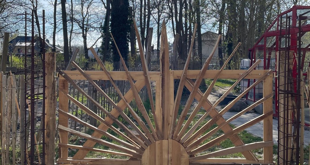 Ein Holztor in Form einer aufgehenden Sonne mit strahlenden Balken steht am Eingang zu einem Garten und wirft dramatische Schatten auf das Gras und den Weg darunter. Im Hintergrund sind kahle Bäume und der blaue Himmel zu sehen.