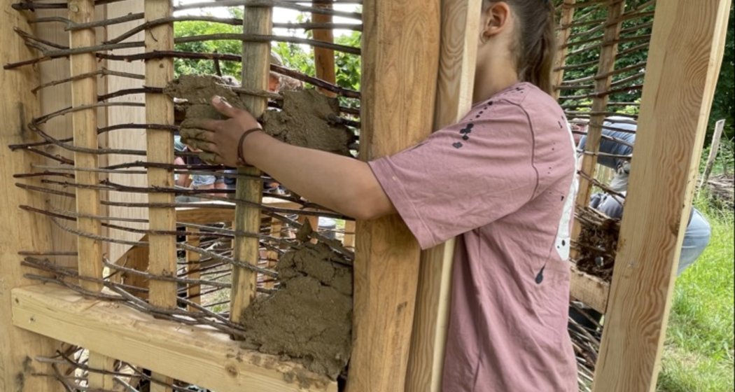 Eine Person in rosa Hemd und Jeans trägt im Freien Schlamm auf eine Struktur aus Holz und geflochtenen Stöcken auf, möglicherweise als Teil eines natürlichen Bauprojekts.