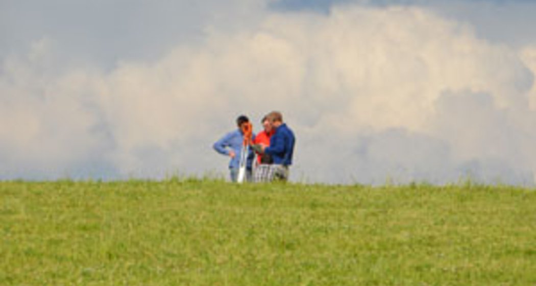 Drei Personen stehen auf einem grasbewachsenen Hügel unter einem wolkenverhangenen Himmel und führen ein Gespräch. Die Szene ist friedlich und offen, wobei das grüne Gras den Vordergrund dominiert.
