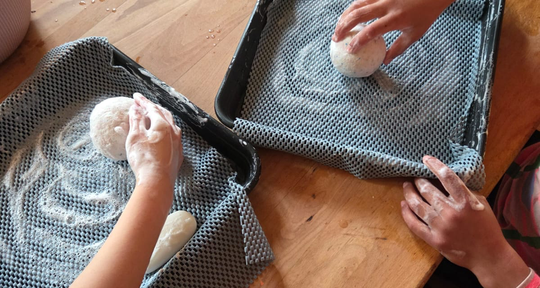 Zwei Kinder formen mit ihren Händen weiße Teigkugeln auf mit Tüchern ausgelegten Tabletts. Auf einem Holztisch befinden sich weitere Tabletts, Tücher und eine Schüssel, was auf eine praktische Tätigkeit hindeutet, wahrscheinlich Brotbacken oder Backen.