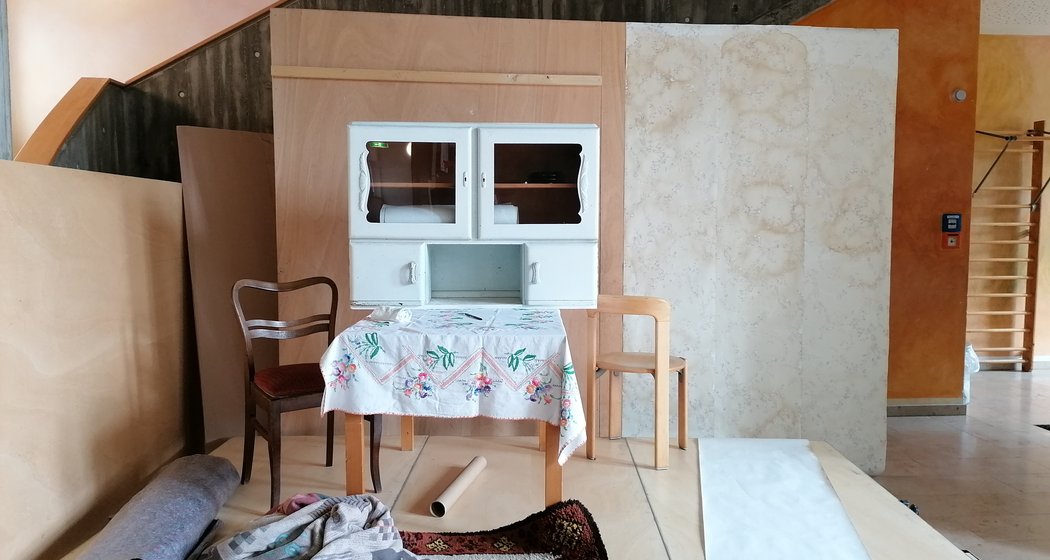 Ein kleiner inszenierter Raum mit einem alten Schrank, zwei nicht zusammenpassenden Stühlen, einem mit einem Blumentuch bedeckten Tisch und einem gemusterten Teppich auf dem Boden, vor unfertigen Wänden und einer Treppe.