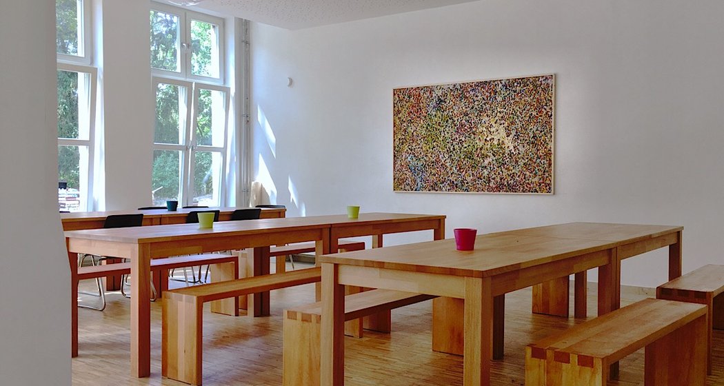 Ein heller Raum mit großen Fenstern, Holztischen und -bänken, ein paar bunten Tassen auf den Tischen und einem abstrakten Gemälde an der weißen Wand. Das Sonnenlicht strömt herein und schafft eine einladende Atmosphäre.