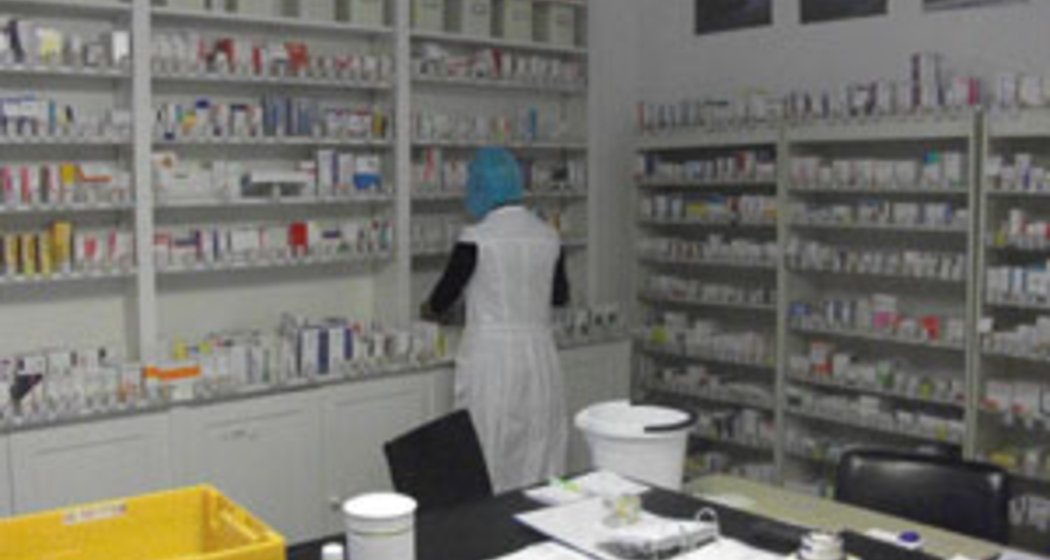 Eine Person in einem weißen Laborkittel und mit blauem Haarschmuck steht in einer Apotheke und ordnet Regale, die mit verschiedenen Medikamentenschachteln und Flaschen gefüllt sind. Ein Tisch im Vordergrund ist mit Papieren und Apothekenbedarf bedeckt.