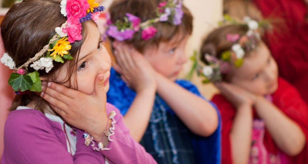 Drei kleine Kinder, die bunte Blumenkronen und Kostüme tragen, stehen in einer Reihe und haben jeweils die Hände an die Wangen gepresst. Es scheint, als würden sie gemeinsam in einer spielerischen oder festlichen Umgebung posieren oder etwas vorführen.