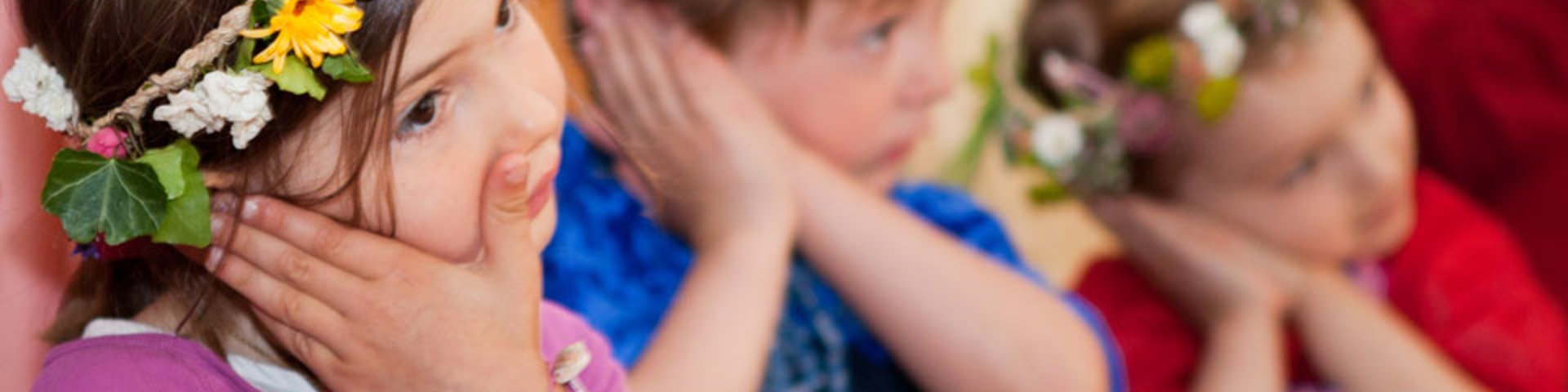 Drei kleine Kinder, die bunte Blumenkronen und Kostüme tragen, stehen in einer Reihe und haben jeweils die Hände an die Wangen gepresst. Es scheint, als würden sie gemeinsam in einer spielerischen oder festlichen Umgebung posieren oder etwas vorführen.