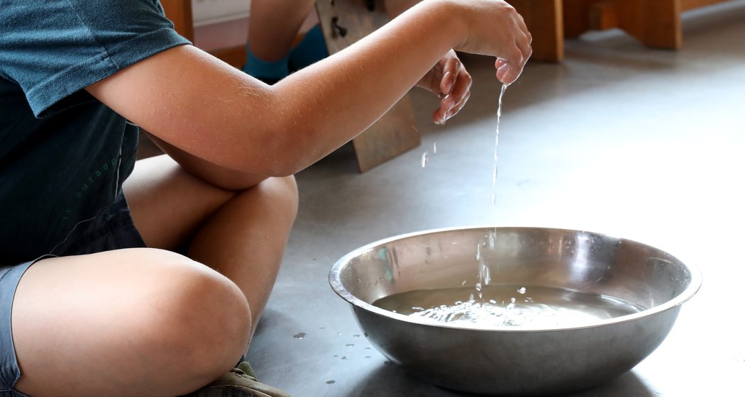 Ein Kind sitzt auf dem Boden und tröpfelt mit einer Hand Wasser in eine große Metallschüssel, während eine andere Person im Hintergrund teilweise sichtbar ist. Die Szene spielt sich in einem Innenraum auf einem grauen Boden ab.