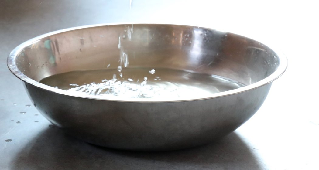 Ein Kind sitzt auf dem Boden und tröpfelt mit einer Hand Wasser in eine große Metallschüssel, während eine andere Person im Hintergrund teilweise sichtbar ist. Die Szene spielt sich in einem Innenraum auf einem grauen Boden ab.