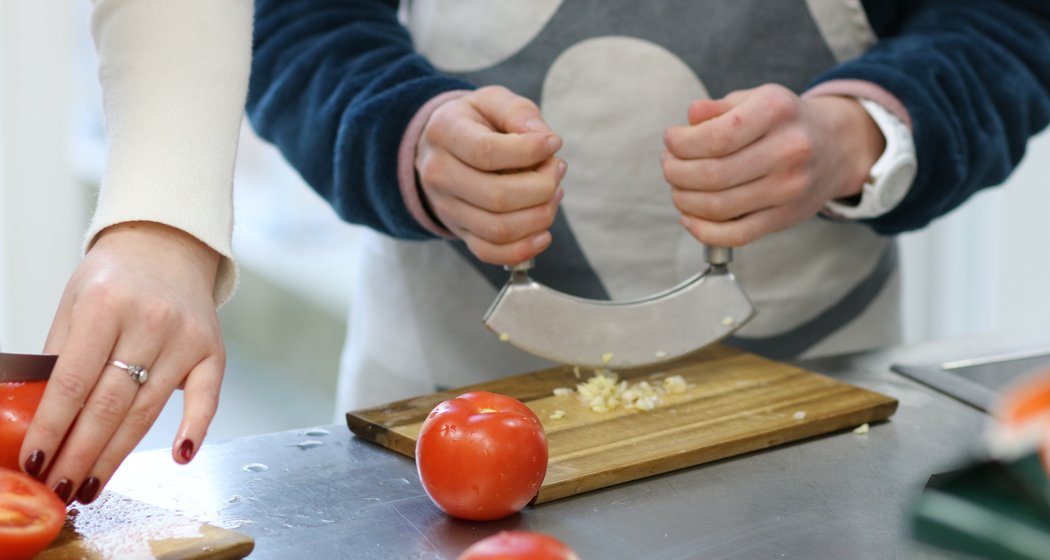 Zwei Personen bereiten auf einem Schneidebrett Lebensmittel zu: die eine hackt Knoblauch mit einem gebogenen Messer, die andere schneidet eine Tomate. Beide sind auf ihre Arbeit konzentriert, und auf der Theke sind frische Tomaten zu sehen.