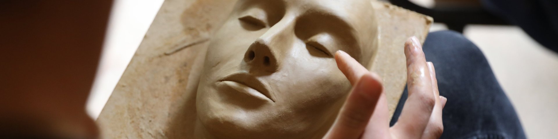 Eine Person formt ein realistisches menschliches Gesicht aus Ton, indem sie die Gesichtszüge sanft mit den Fingern formt, während sie das Kunstwerk auf ihrem Schoß hält.
