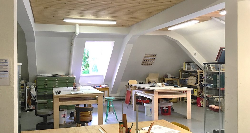 Ein helles Kunstatelier mit Holztischen, Hockern, Kunstzubehör und mit Materialien gefüllten Regalen. Schräge Decke mit Holzverkleidung, große Fenster und natürliches Licht erhellen den Arbeitsbereich.
