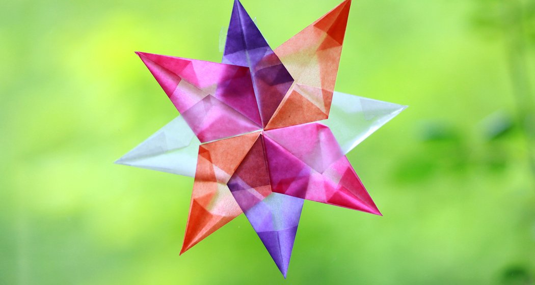 Ein farbenfroher, achtzackiger Origami-Stern mit sich überlappenden, durchscheinenden Segmenten in Rot, Lila, Orange und Weiß vor einem verschwommenen grünen Hintergrund.