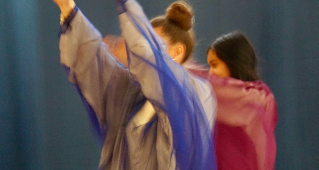 Zwei Personen tanzen mit erhobenen Armen in fließenden, durchscheinenden Gewändern - eine in Blau und eine in Rosa - vor einem verschwommenen, dunklen Hintergrund, der Bewegung und Freude suggeriert.