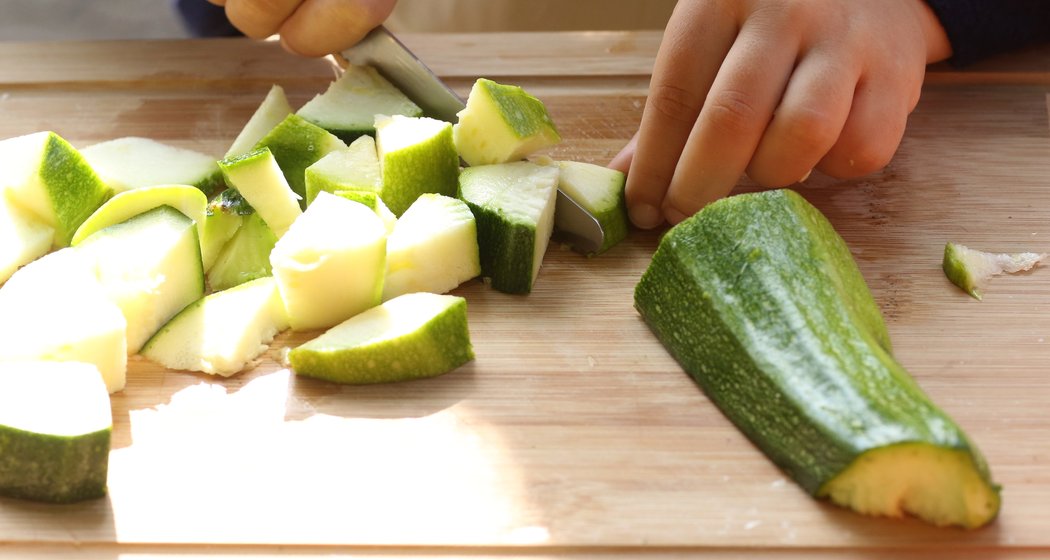 Eine Person schneidet grüne Zucchini mit einem Messer auf einem Holzbrett in Würfel, wobei eine Hand das Gemüse hält und die andere schneidet.