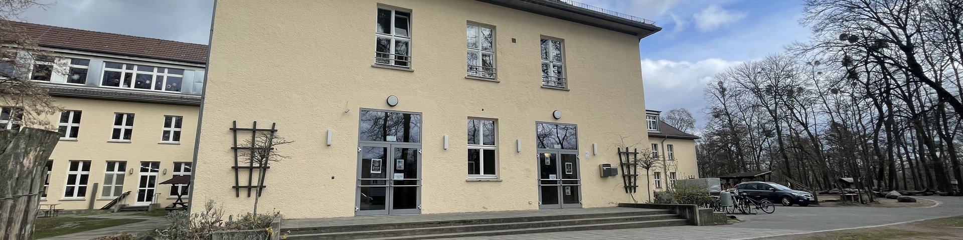 Ein großes beigefarbenes Gebäude mit mehreren Fenstern und doppelten Glastüren steht unter einem wolkenverhangenen Himmel, umgeben von kahlen Bäumen und einer Grasfläche mit einem großen Felsbrocken im Vordergrund.