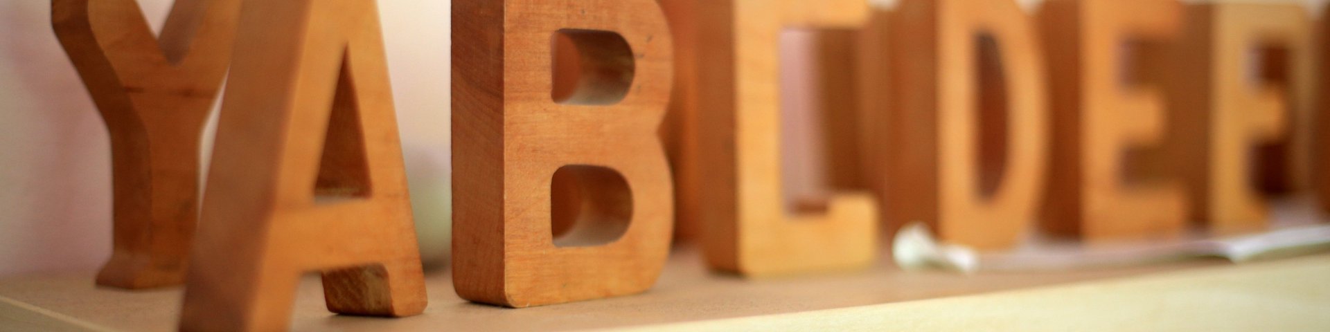 Holzblockbuchstaben, die YABCD und DEE schreiben, sind auf einem hellen Holzregal vor einem blassen Hintergrund angeordnet. Der Fokus liegt auf den Buchstaben im Vordergrund, während andere leicht verschwommen sind.