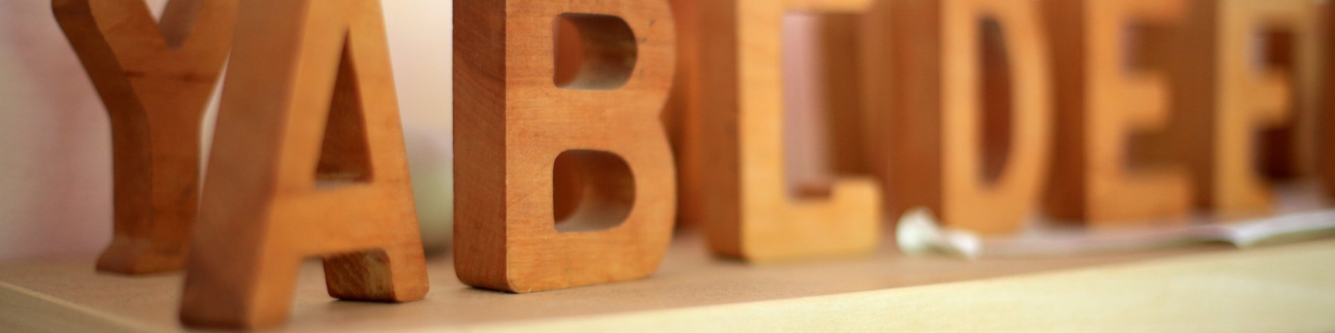 Holzblockbuchstaben, die YABCD und DEE schreiben, sind auf einem hellen Holzregal vor einem blassen Hintergrund angeordnet. Der Fokus liegt auf den Buchstaben im Vordergrund, während andere leicht verschwommen sind.