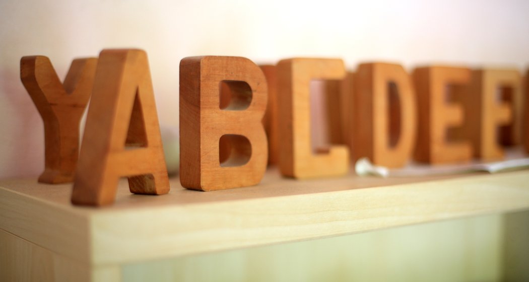 Holzblockbuchstaben, die YABCD und DEE schreiben, sind auf einem hellen Holzregal vor einem blassen Hintergrund angeordnet. Der Fokus liegt auf den Buchstaben im Vordergrund, während andere leicht verschwommen sind.