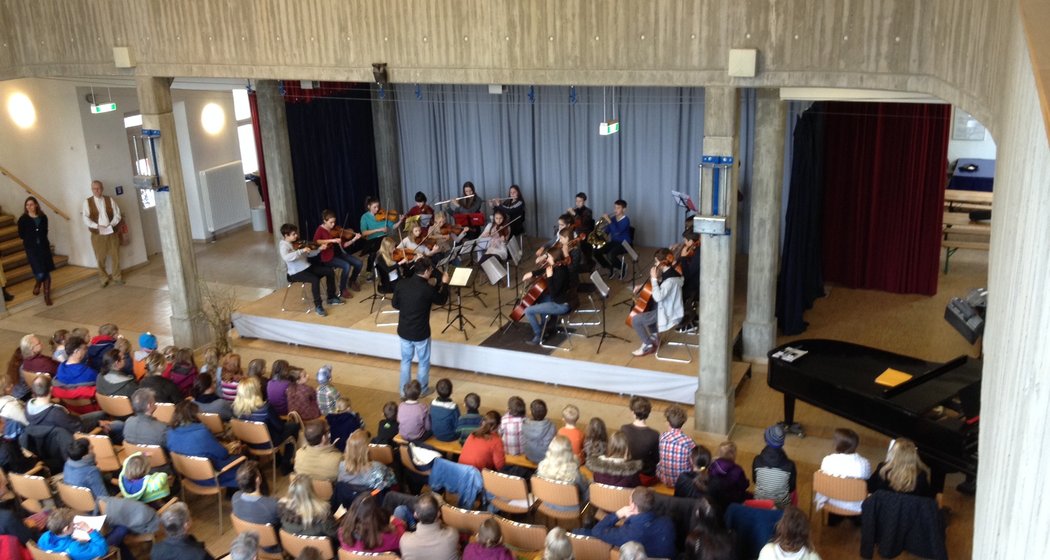Blick von oben auf ein kleines Orchester, das in einem Saal auf der Bühne spielt, während das Publikum sitzt und zusieht. Ein Dirigent steht vor den Musikern, und rechts von der Bühne ist ein Flügel zu sehen.
