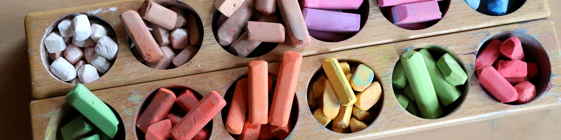 Ein Holztablett mit runden Fächern, die mit bunten Kreidestücken gefüllt sind, darunter weiße, braune, rosafarbene, rote, orangefarbene, gelbe, grüne und blaue, die auf einer hellen Holzoberfläche platziert sind.
