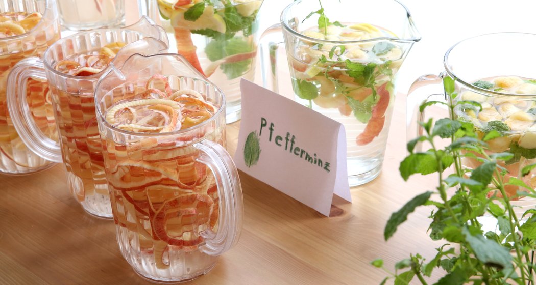 Ein Tisch mit Krügen und Gläsern mit Fruchtwasser, das Zitrone, Orange und Minze enthält. Eine handgeschriebene Karte mit der Aufschrift "Pfefferminze" liegt in der Nähe, neben einer frischen Minzpflanze.