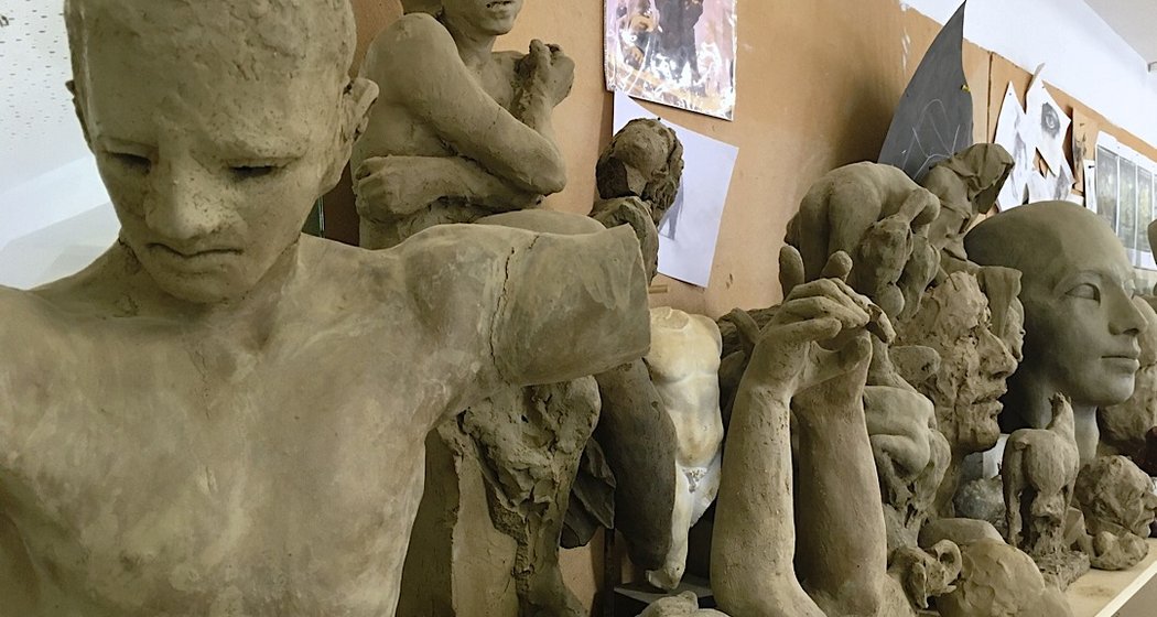 Eine Reihe unvollendeter Tonskulpturen, die menschliche Figuren und Gesichter darstellen, stehen dicht nebeneinander auf einem Regal in einem Kunstatelier. An der Wand dahinter hängen verschiedene Skizzen und Kunstmaterialien.