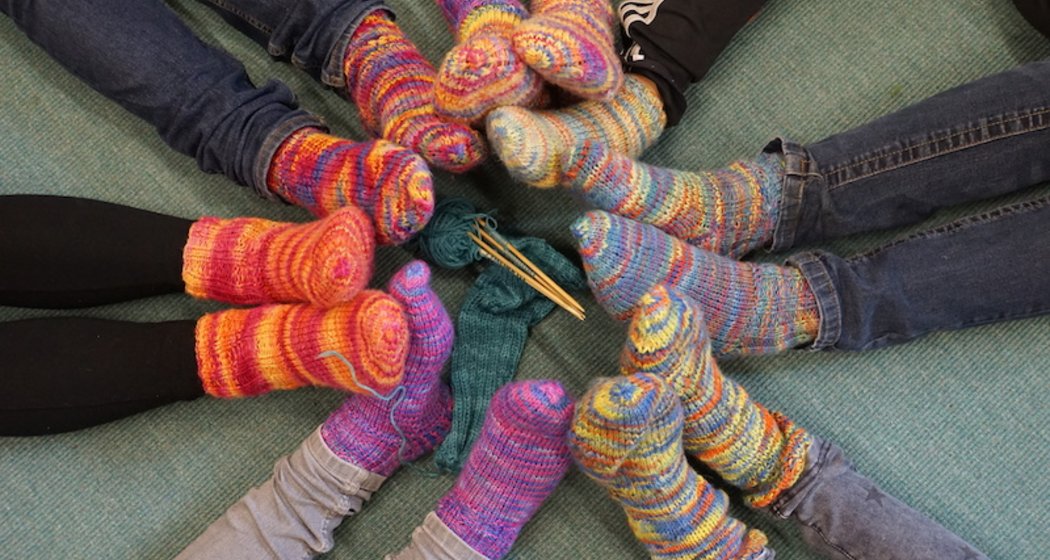 Eine Gruppe von Menschen sitzt in einem Kreis auf einem grünen Teppich, jeder trägt bunte, gemusterte Stricksocken. Die Füße sind in die Mitte gerichtet, wo Stricknadeln und Garn platziert sind.