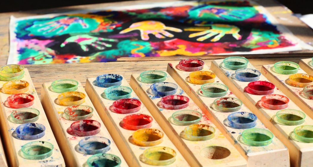 Bunte Aquarellfarben liegen auf einem Holztisch, mit einem lebendigen Handabdruckbild im Hintergrund. Das Bild zeigt mehrere helle Handabdrücke und Farbspritzer auf Papier.