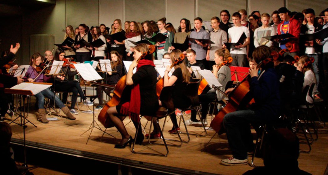 Ein Chor von Jugendlichen steht auf Steigbügeln und singt, während ein sitzendes Orchester von jungen Musikern auf einer Bühne Streichinstrumente spielt. Ein Dirigent steht links und leitet die Gruppe. Es scheint sich um ein Hallenkonzert zu handeln.